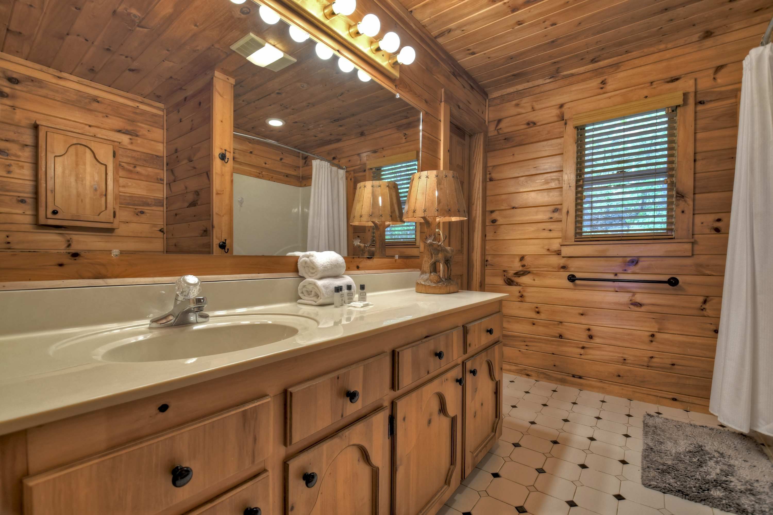 A wooden cabin bathroom with a large mirror, a sink, and a window with blinds.