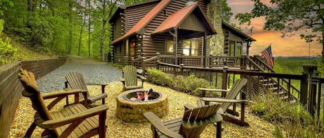 A log cabin with a stone chimney, outdoor seating with Adirondack chairs, and a fire pit.