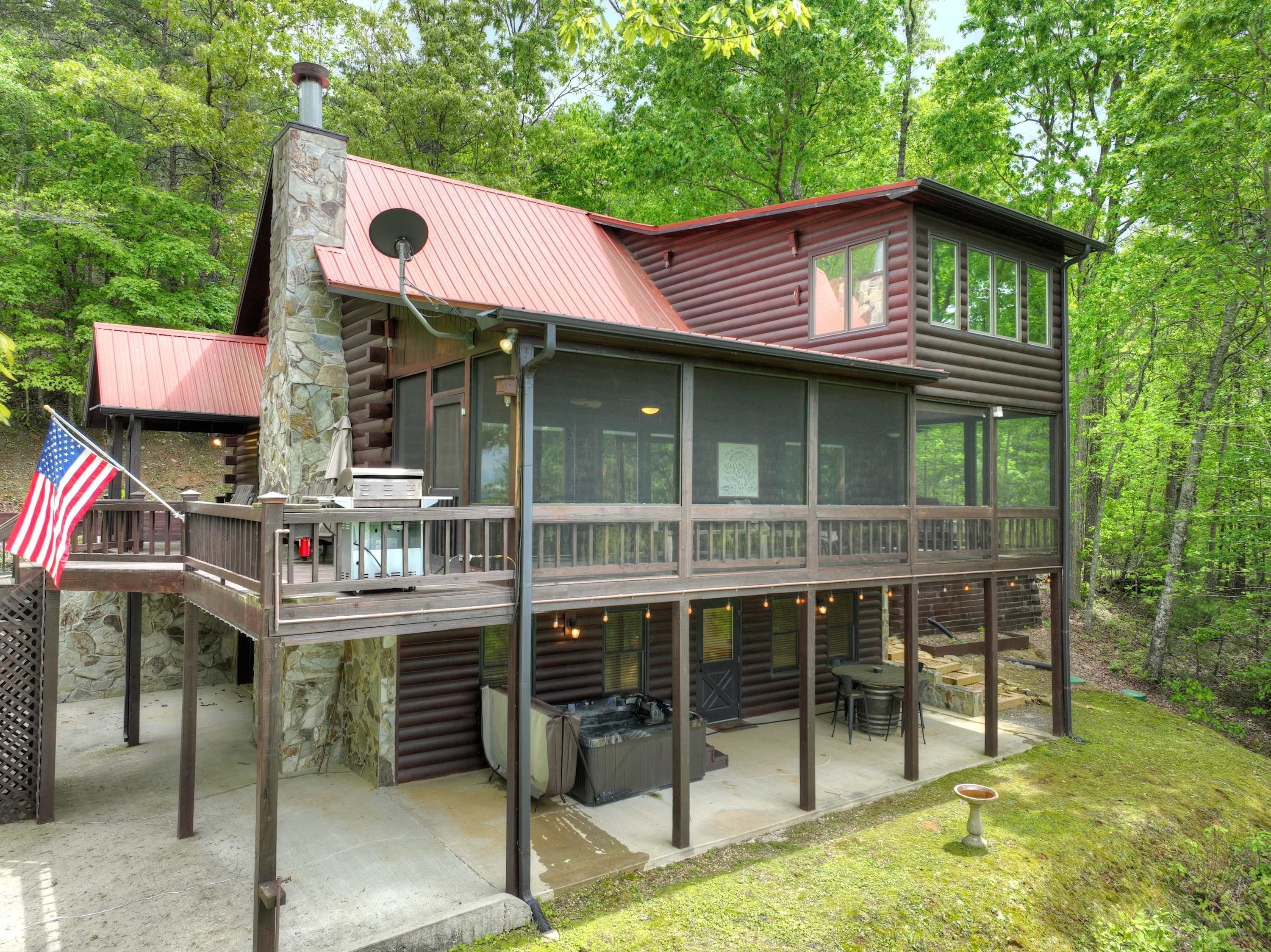 A two-story log cabin with a stone chimney, a deck with outdoor seating, and an American flag.