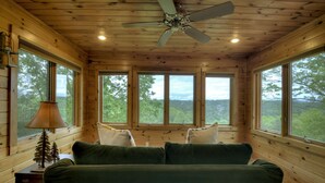 Ein Holzblockhaus-Interieur mit einem grünen Sofa, einem Deckenventilator und großen Fenstern mit Blick auf den Wald.