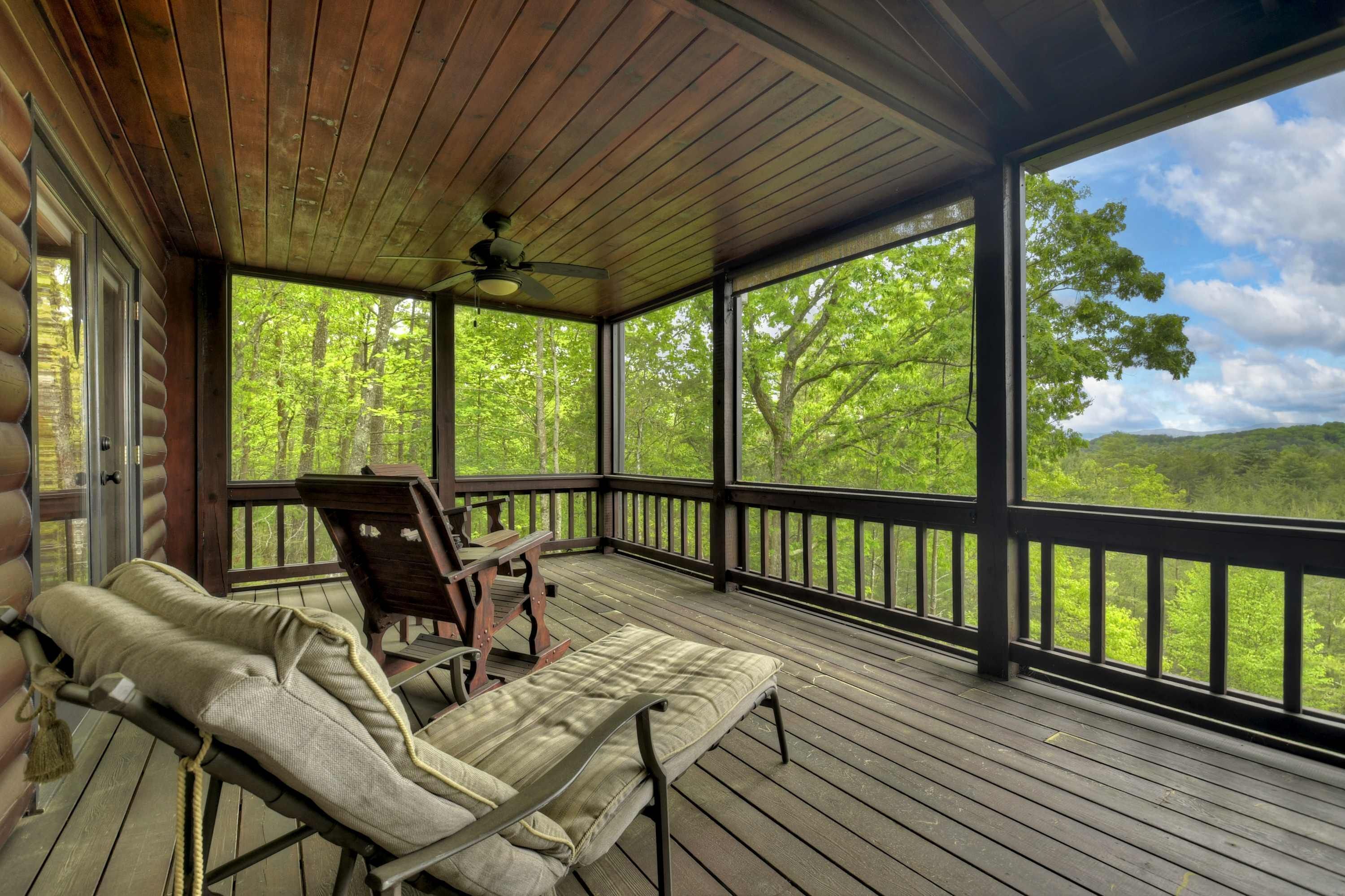 A covered porch with a ceiling fan, wooden deck, and a view of a forested landscape.