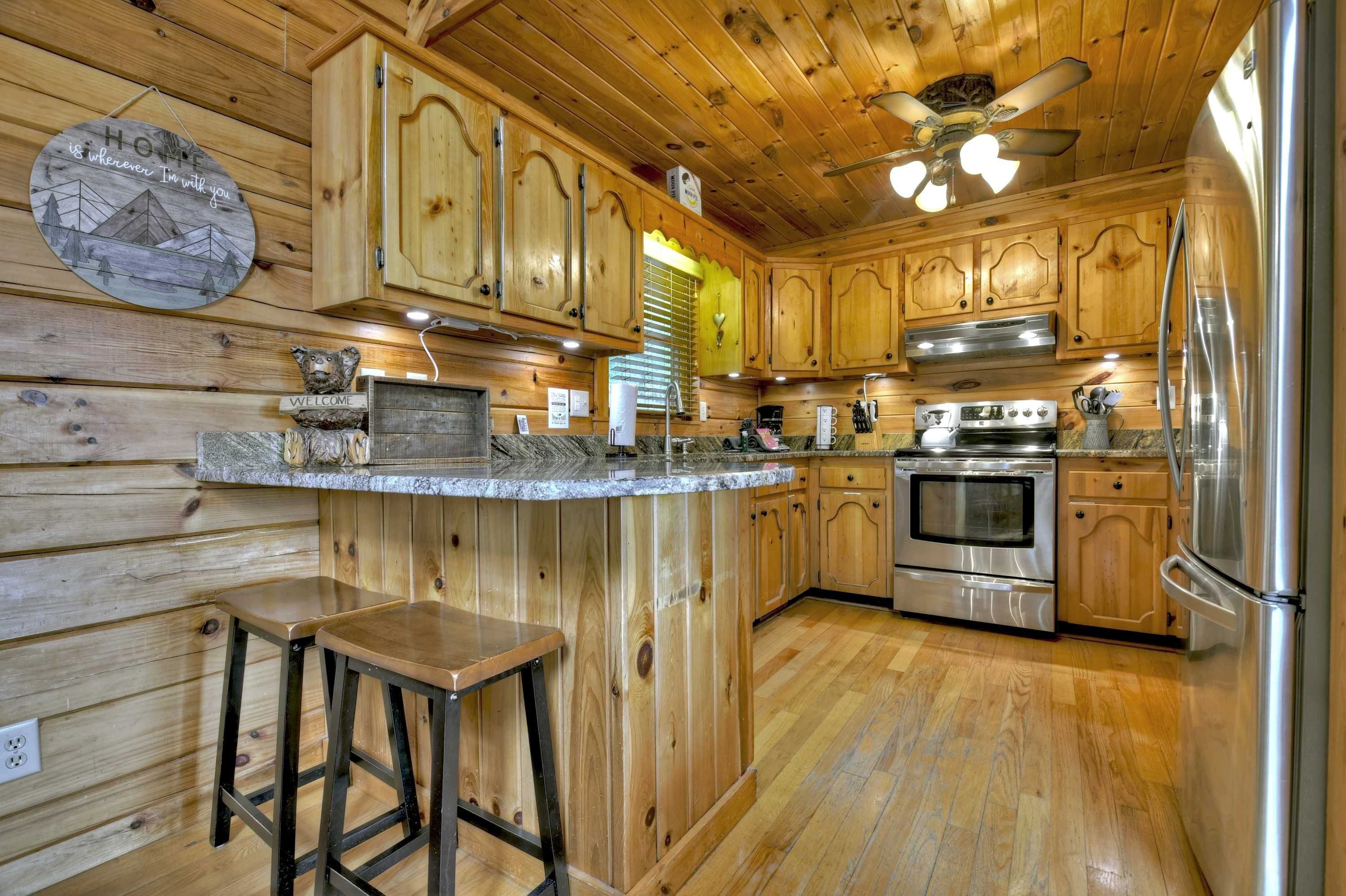 A wooden cabin interior with a kitchen featuring wooden cabinets, a central island with stools, a ceiling fan, and stainless steel appliances.