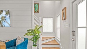 Una escalera de madera con peldaños que lleva a una plataforma, una silla azul con cojín, una planta en maceta y dos cuadros en la pared.