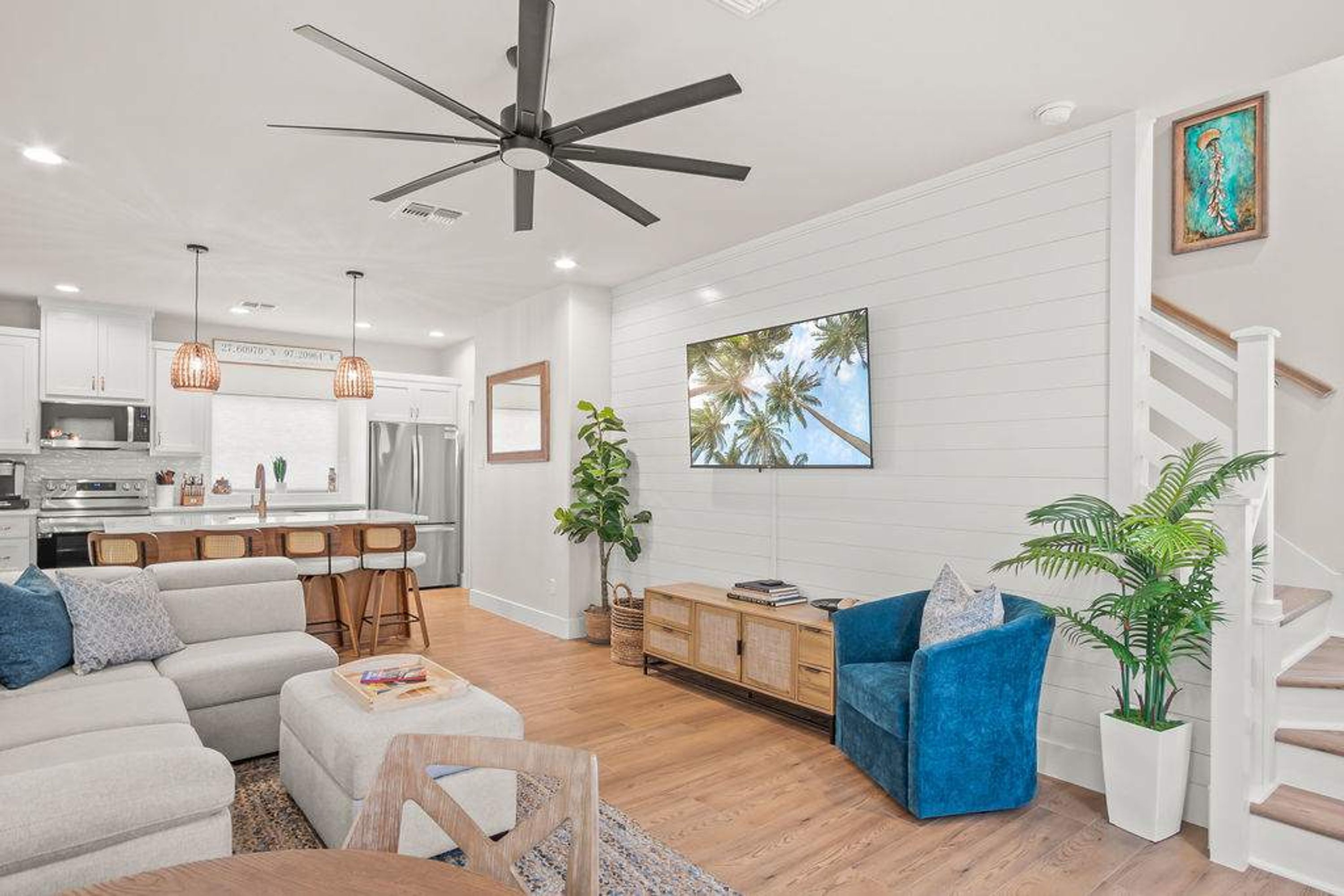 A modern living room with a large ceiling fan, a kitchen with white cabinets, and a dining area with wooden chairs.