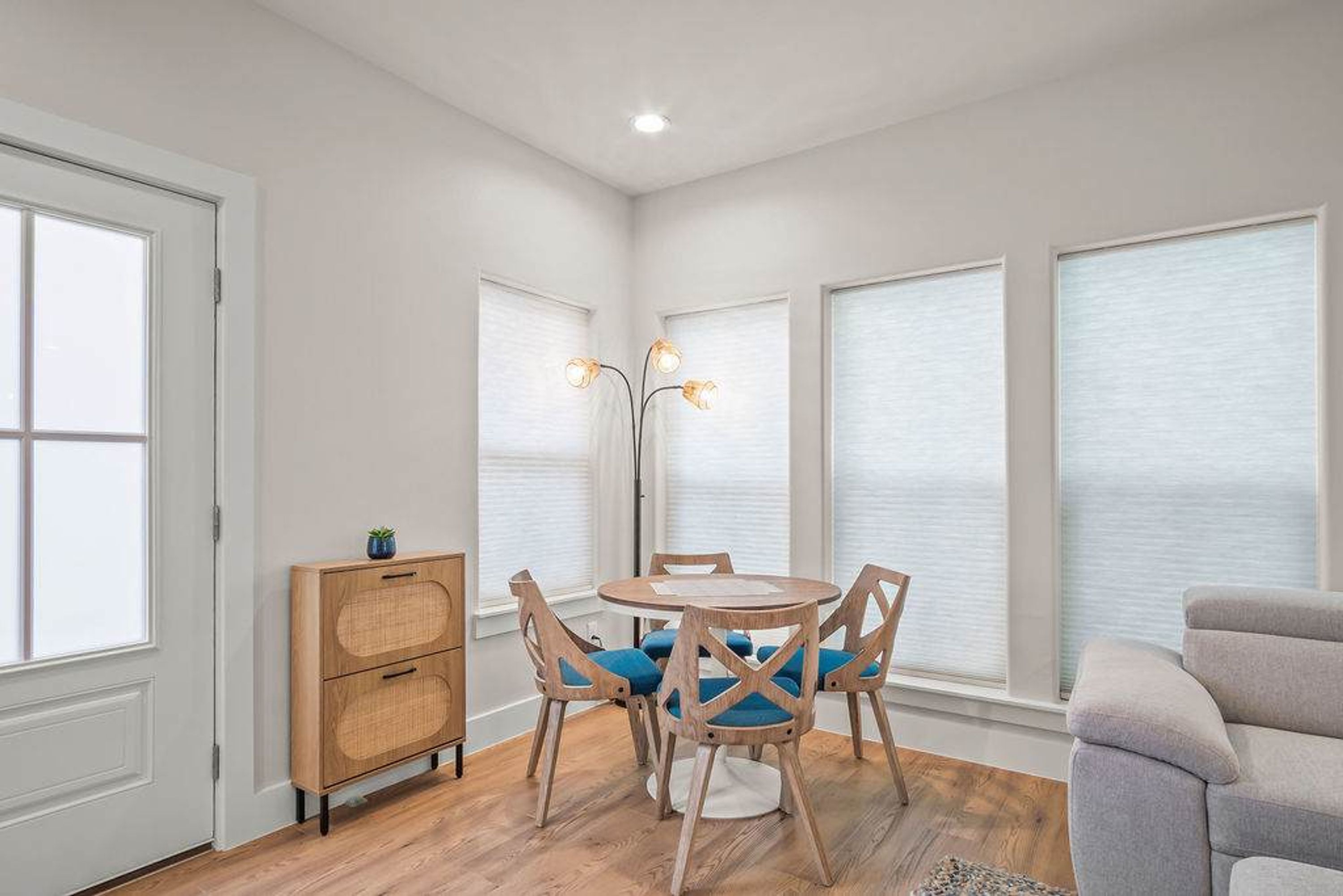 A room with a round wooden table, four chairs, a floor lamp, a small cabinet, and a grey sofa.