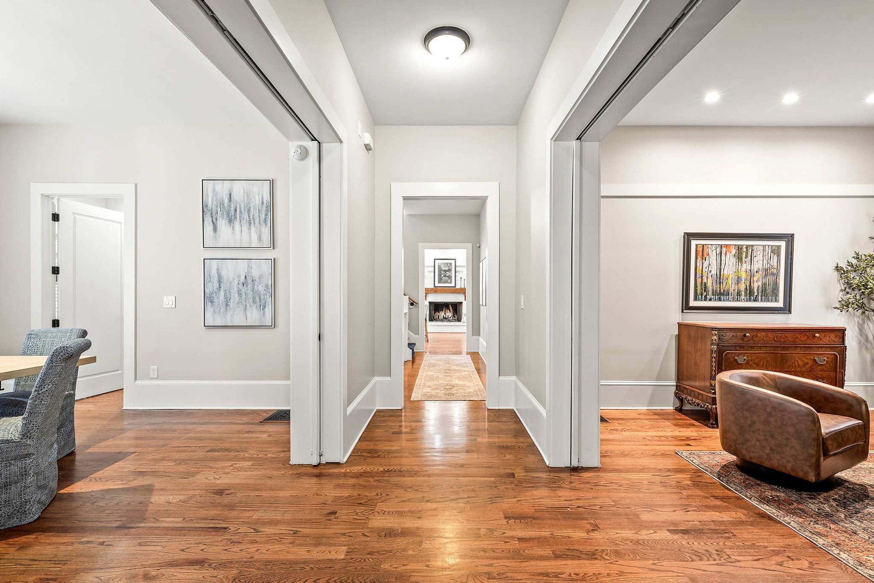 A hallway with hardwood flooring, white walls, and a fireplace in the distance. There are framed artworks on the walls and a brown leather armchair.