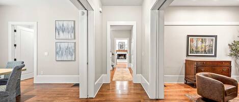 A hallway with hardwood flooring, white walls, and a fireplace in the distance. There are framed artworks on the walls and a brown leather armchair.