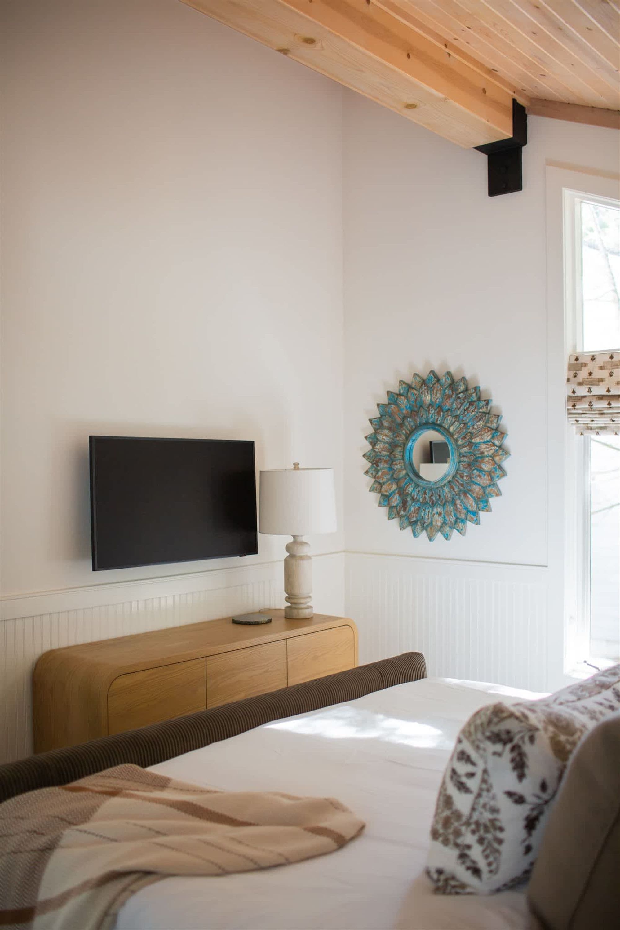 Quarto com cama, televisão, abajur e um espelho decorativo na parede.