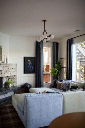 Ein modernes Wohnzimmer mit Kamin aus Stein, einer Sofagarnitur, einem großen Fenster mit Vorhängen und einer Pendelleuchte.