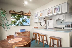 Eine kompakte Küche mit weißen Schränken, einem Holztisch mit Hocker und einem großen Fenster mit Blick auf eine tropische Landschaft.