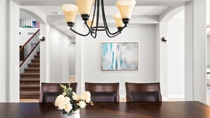 A dining area with a dark wooden table, four chairs, a vase with flowers, a chandelier, and a staircase in the background. - Desert Retreat Pool Fire Pit Family Games (Surprise)