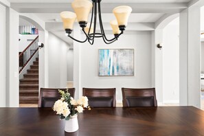 A dining area with a dark wooden table, four chairs, a vase with flowers, a chandelier, and a staircase in the background. - Desert Retreat Pool Fire Pit Family Games (Surprise)