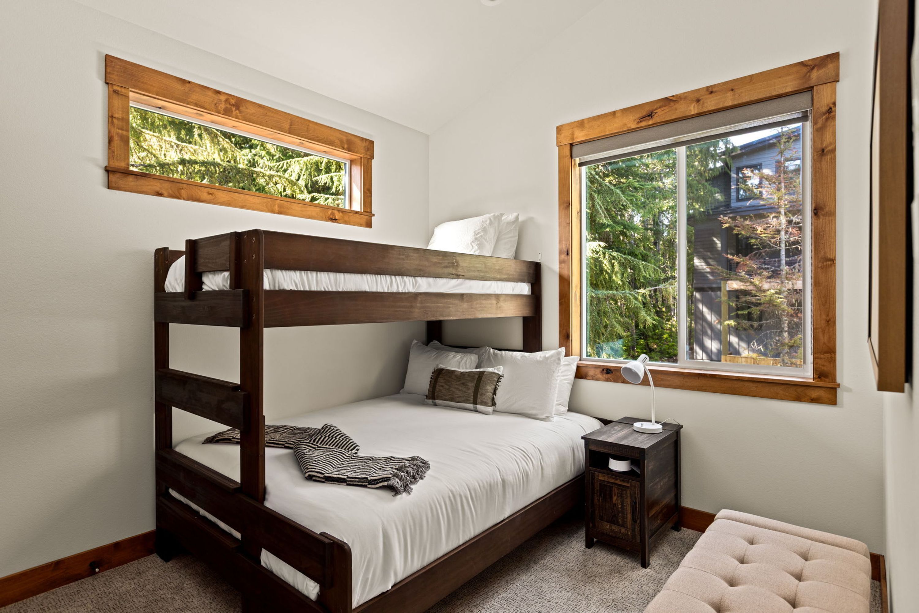 Una cama litera con escritorio y lámpara, una ventana con vista a árboles, y un ventanal pequeño sobre la cama.