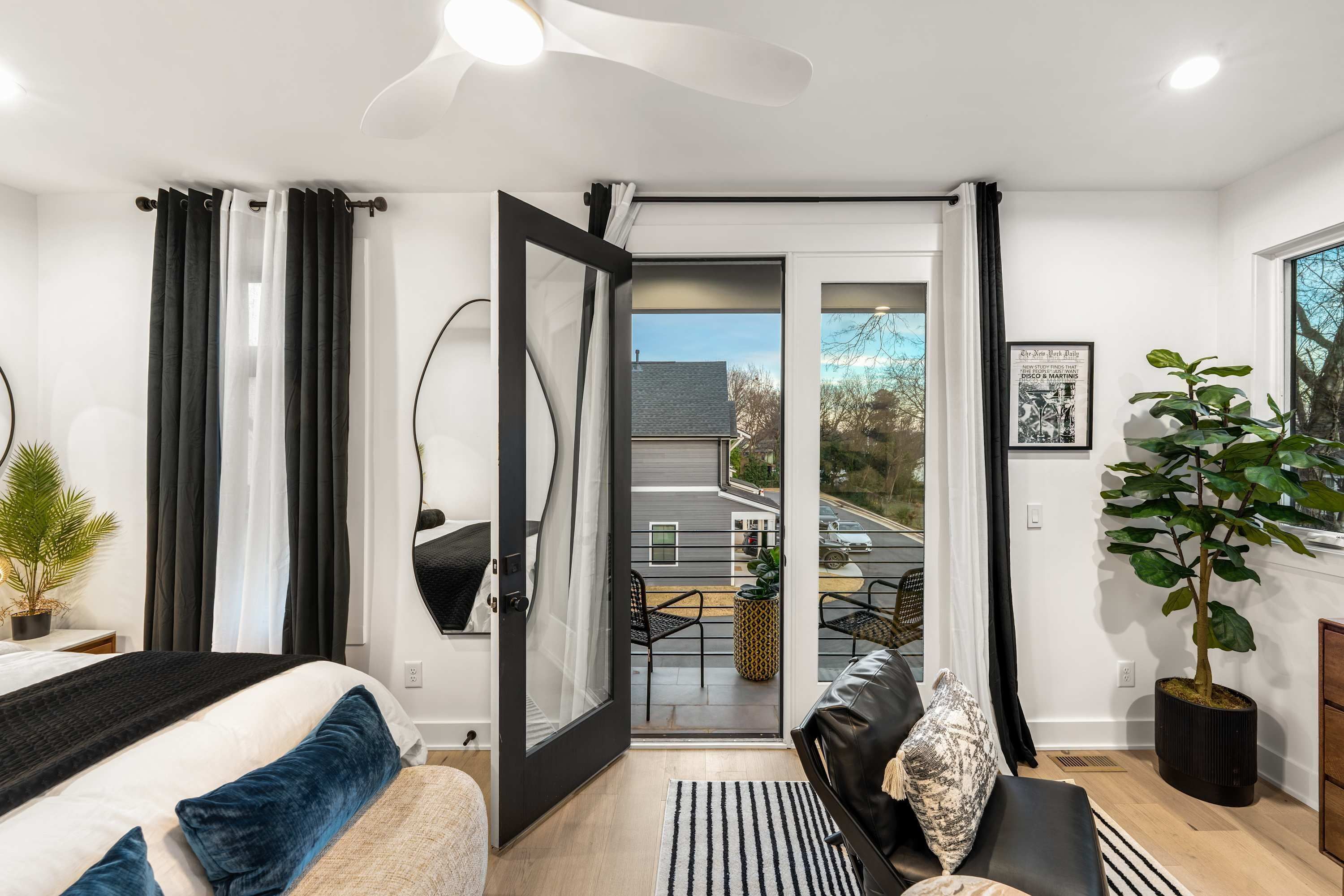 A modern bedroom with a bed, a black-framed mirror, a sliding glass door leading to a balcony, and a potted plant.