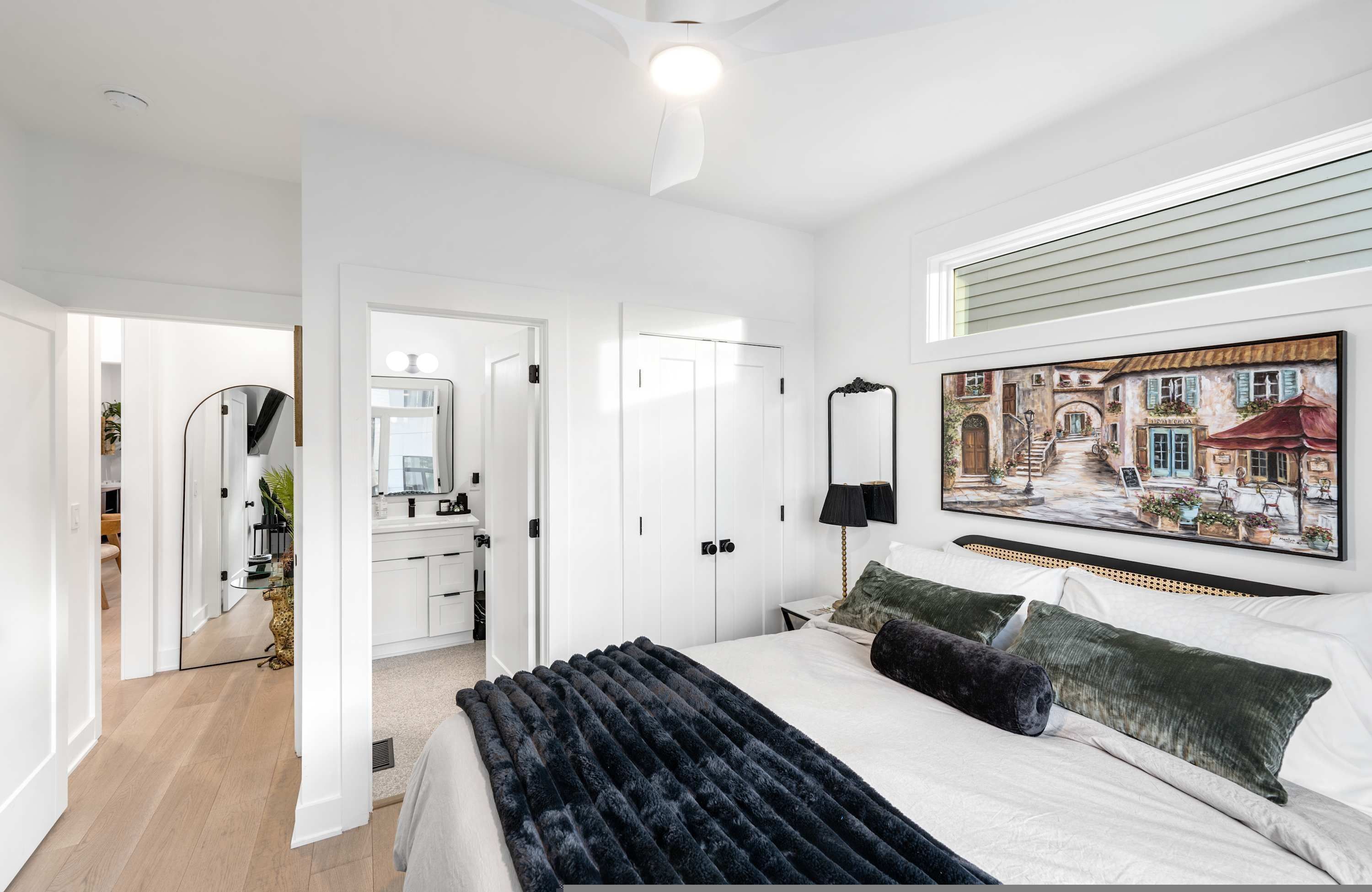 A modern bedroom with a large bed, a painting of a street scene, and a view of a cityscape through the window.