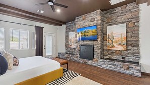 A bedroom with a bed, a bench, a television mounted on a stone wall, and a fireplace. - Luxury 6BR Retreat - Pool BBQ Theater and Games (Mesa)