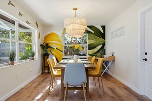 Un espace repas avec une table en bois, des chaises jaunes, un luminaire suspendu et de grandes fenêtres donnant sur un paysage verdoyant.