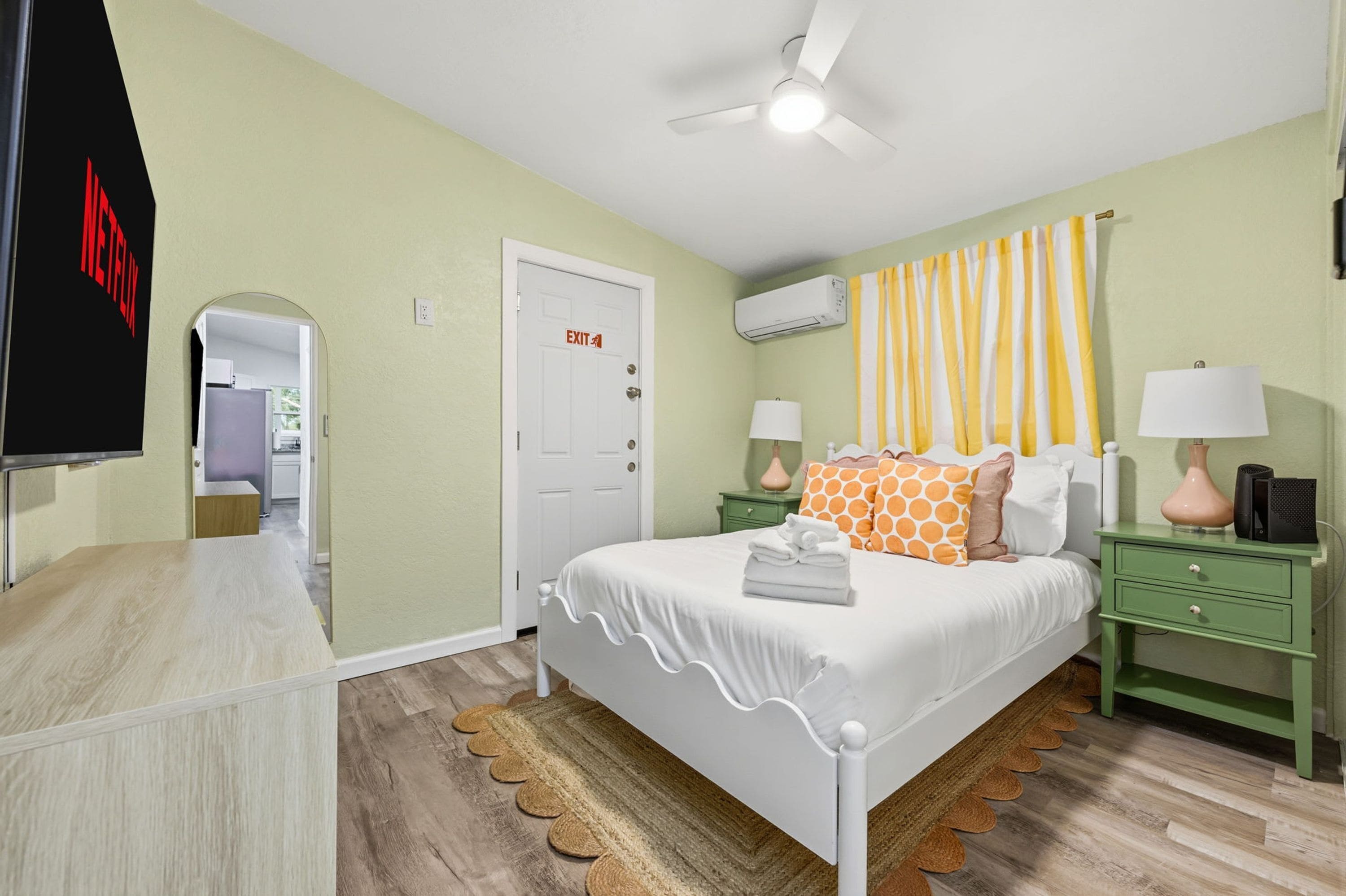 Un dormitorio con cama, mesitas de noche, ventilador de techo, unidad de aire acondicionado y una puerta con señal de salida.