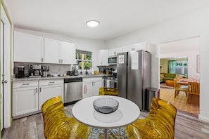 A modern kitchen with white cabinets, stainless steel appliances, and a dining area with yellow chairs and a white table. - Backyard Oasis Heated Pool Tiki Bar Hot Tub (Largo)