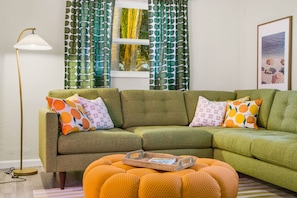 A living room with a green sectional sofa, orange ottoman, patterned pillows, a floor lamp, and a window with green and white patterned curtains. - Backyard Oasis Heated Pool Tiki Bar Hot Tub (Largo)