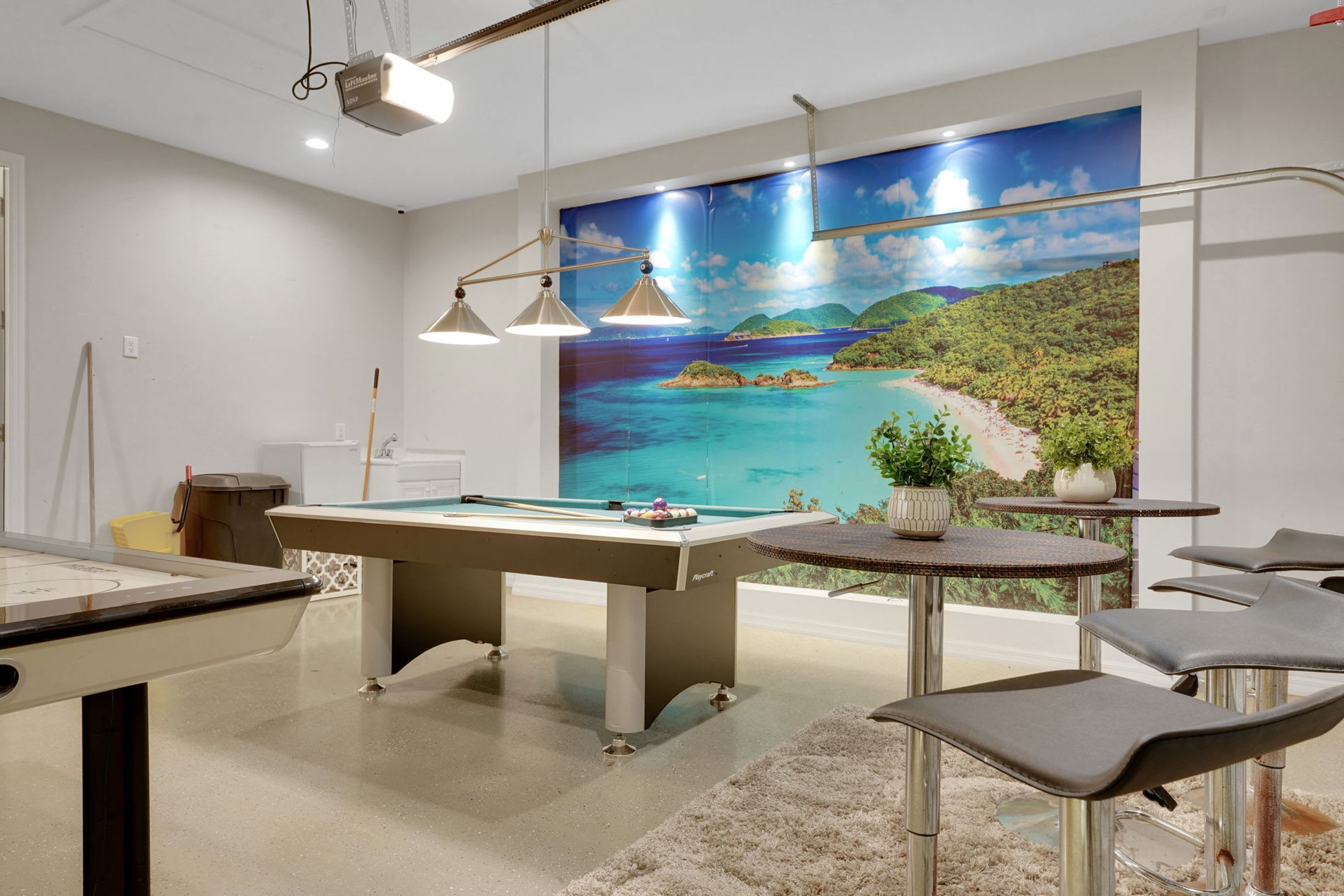 A room with a pool table, a projector, a painting of a beach, and a small table with chairs.