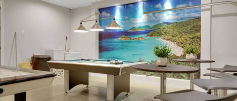 A room with a pool table, a projector, a painting of a beach, and a small table with chairs.