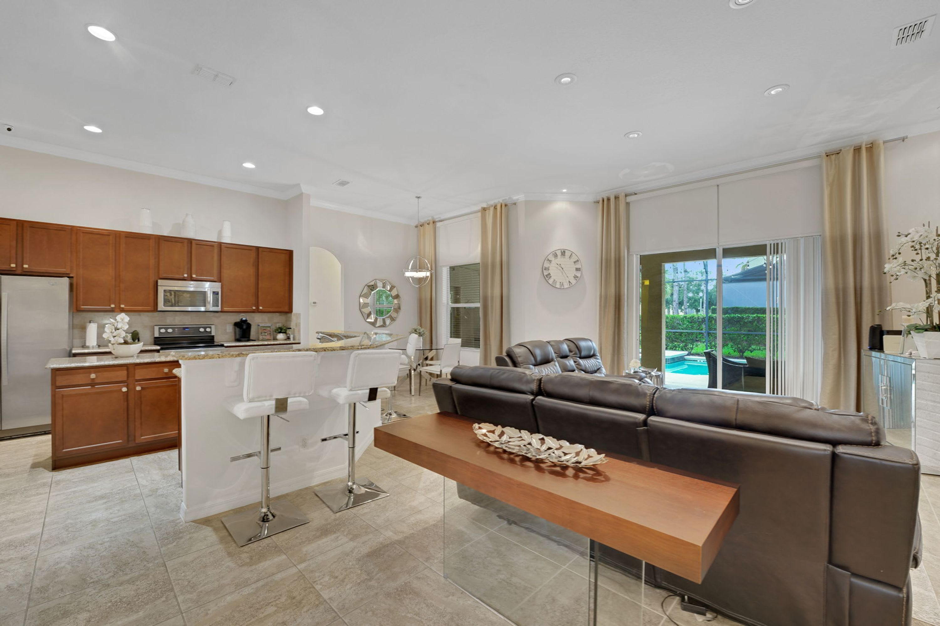 A modern kitchen with a built-in microwave, a dining area with a bar stool, a leather sofa, and a large window with a view of a swimming pool.