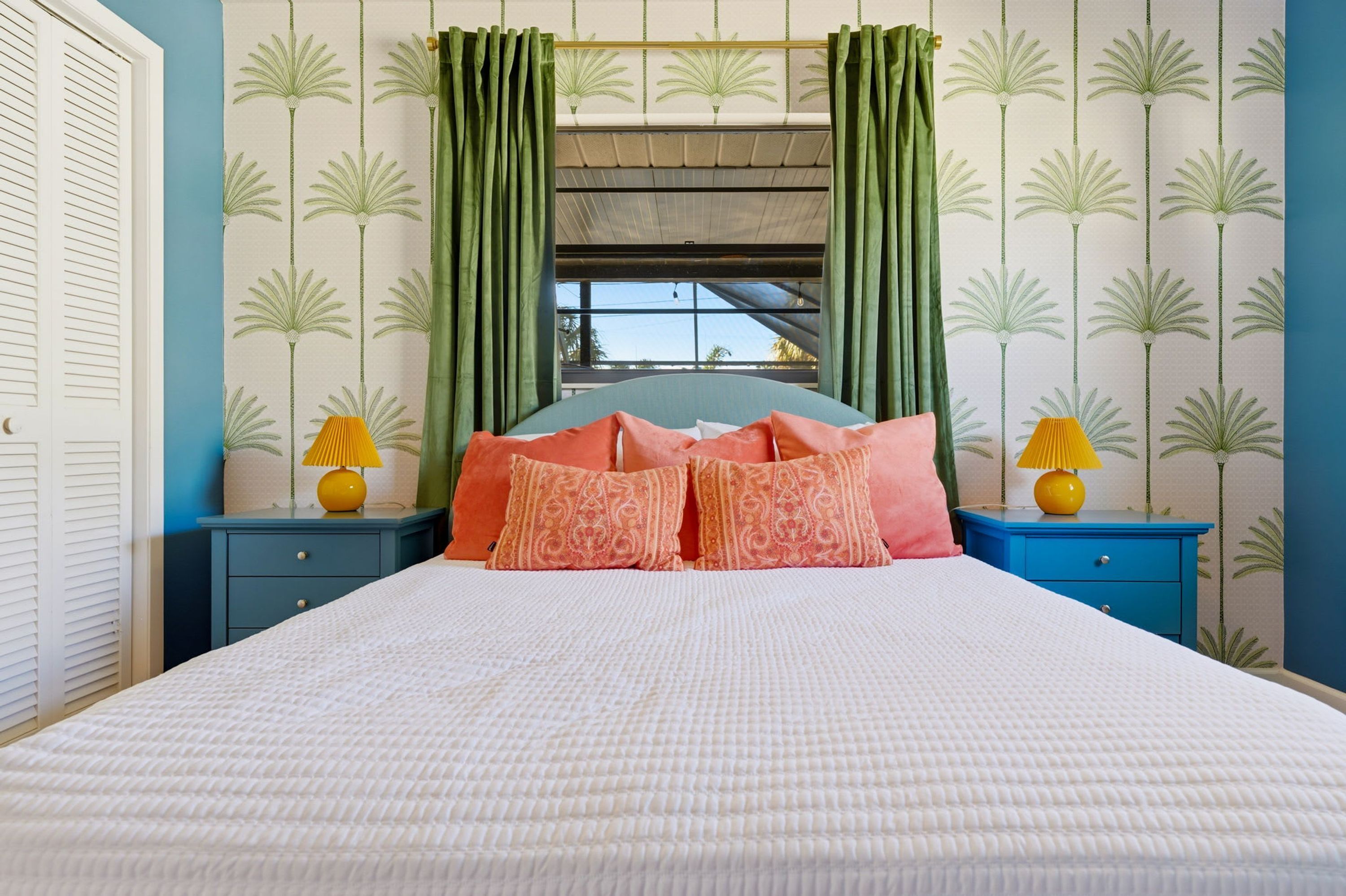 Un dormitorio con una cama, dos mesitas de noche, un ventanal con cortinas y una pared decorada con un estampado de palmeras.