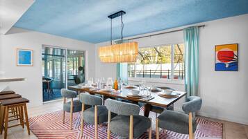 Un comedor con una mesa de madera dispuesta para seis comensales, sillas tapizadas en gris, una lámpara colgante de estilo bambú y vistas a través de grandes ventanales.
