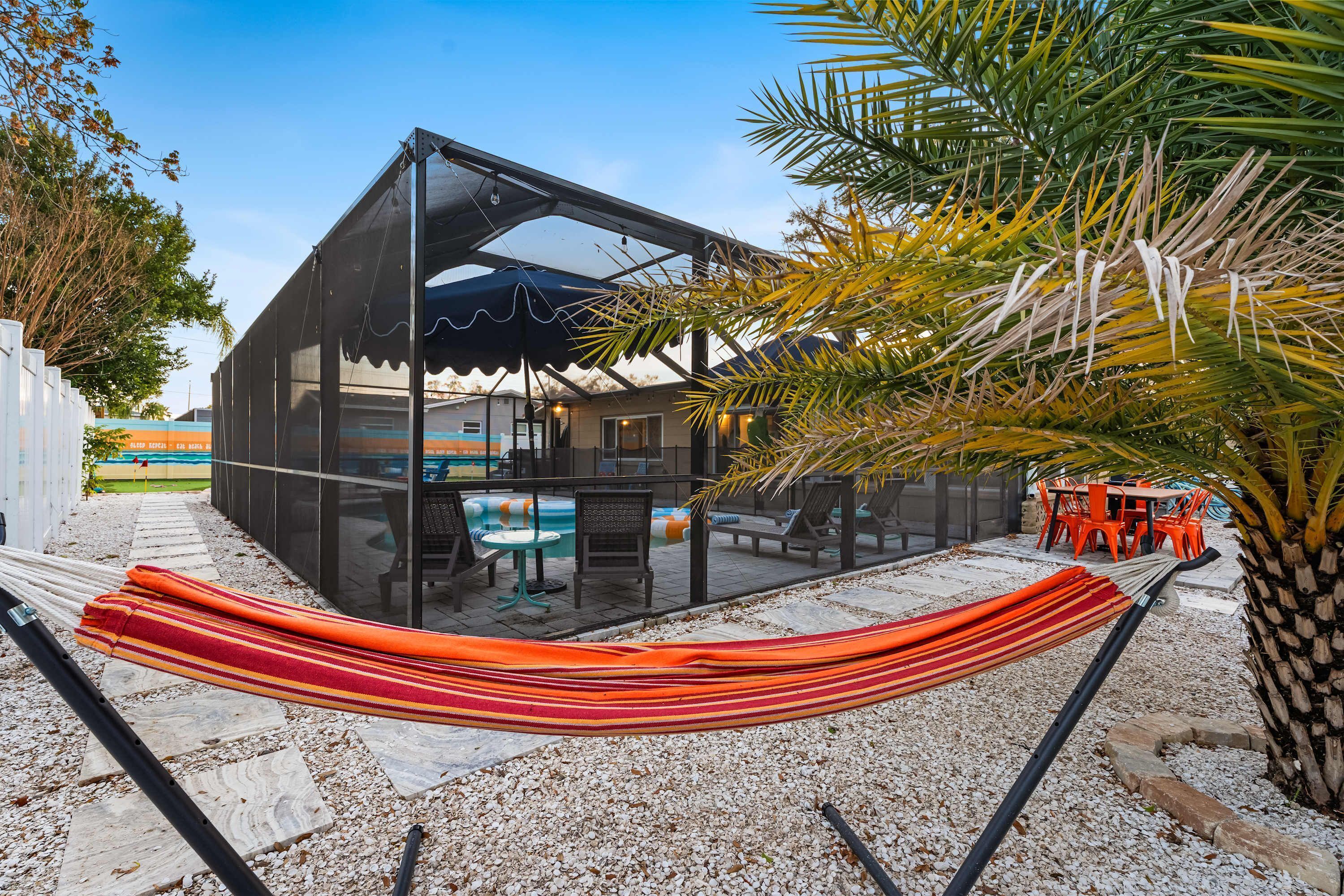 Un área de piscina moderna al aire libre con una hamaca, sillones reclinables y un espacio de descanso techado.