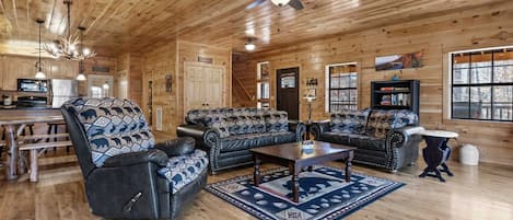 A log cabin interior with a dining area, a living room with leather sofas, a ceiling fan, and a large window.