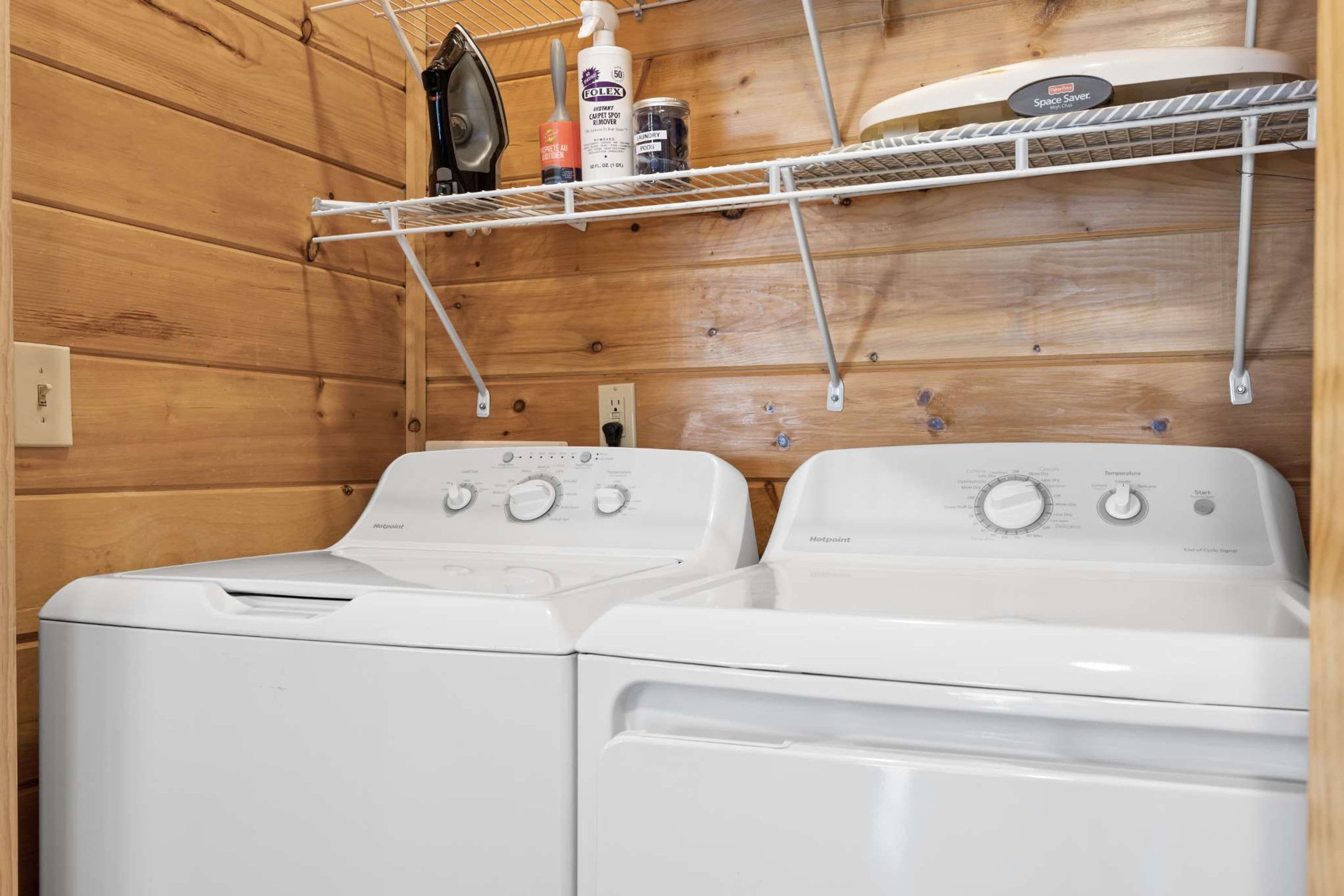 A laundry room with two front-loading washing machines, a shelf with cleaning supplies, and a space saver rack above.