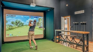 A man playing golf indoors with a large screen displaying a golf course. There is a bar area with stools and a door in the background. - Epic Poconos Pad Pool Hot Tub Gym Golf Sim (East Stroudsburg)