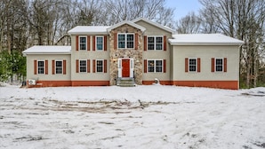 A two-story house with a stone entrance and red shutters, surrounded by snow. - Epic Poconos Pad Pool Hot Tub Gym Golf Sim (East Stroudsburg)