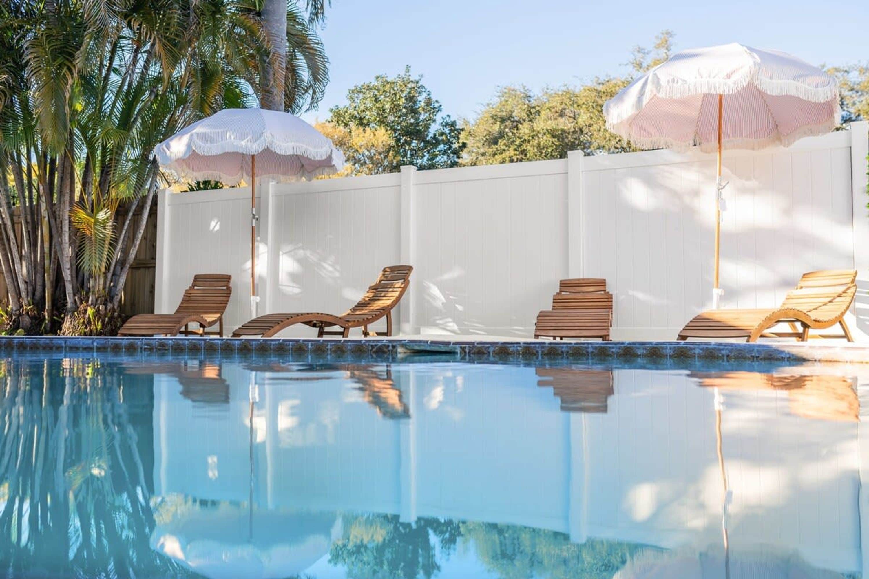 Una piscina con sillas de descanso y sombrillas, rodeada por una cerca blanca y palmeras.