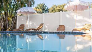 Una piscina con sillas de descanso y sombrillas, rodeada por una cerca blanca y palmeras.