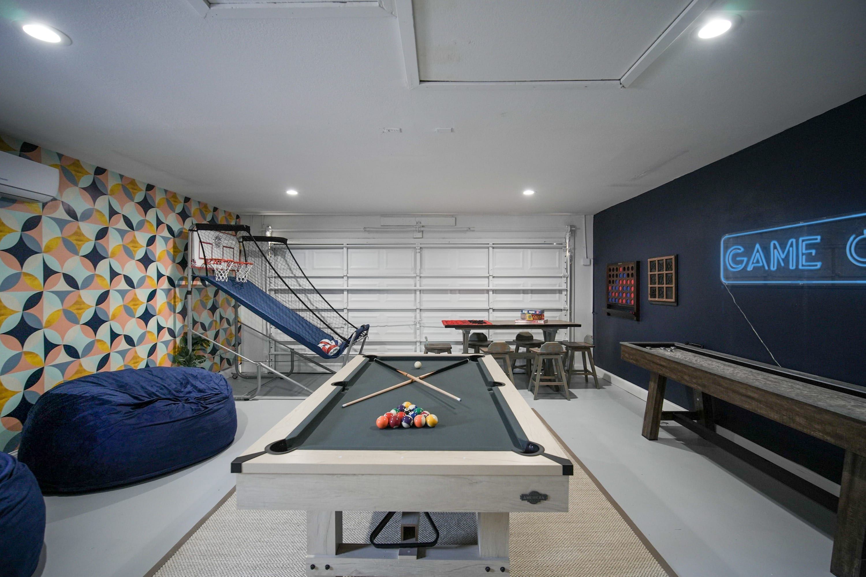 Una mesa de billar con bolas, un sillón tipo bean bag azul, un aro de baloncesto y un letrero de neón que dice 'GAME'.