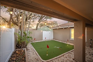 Un patio trasero con un campo de práctica de golf, una valla blanca y árboles.