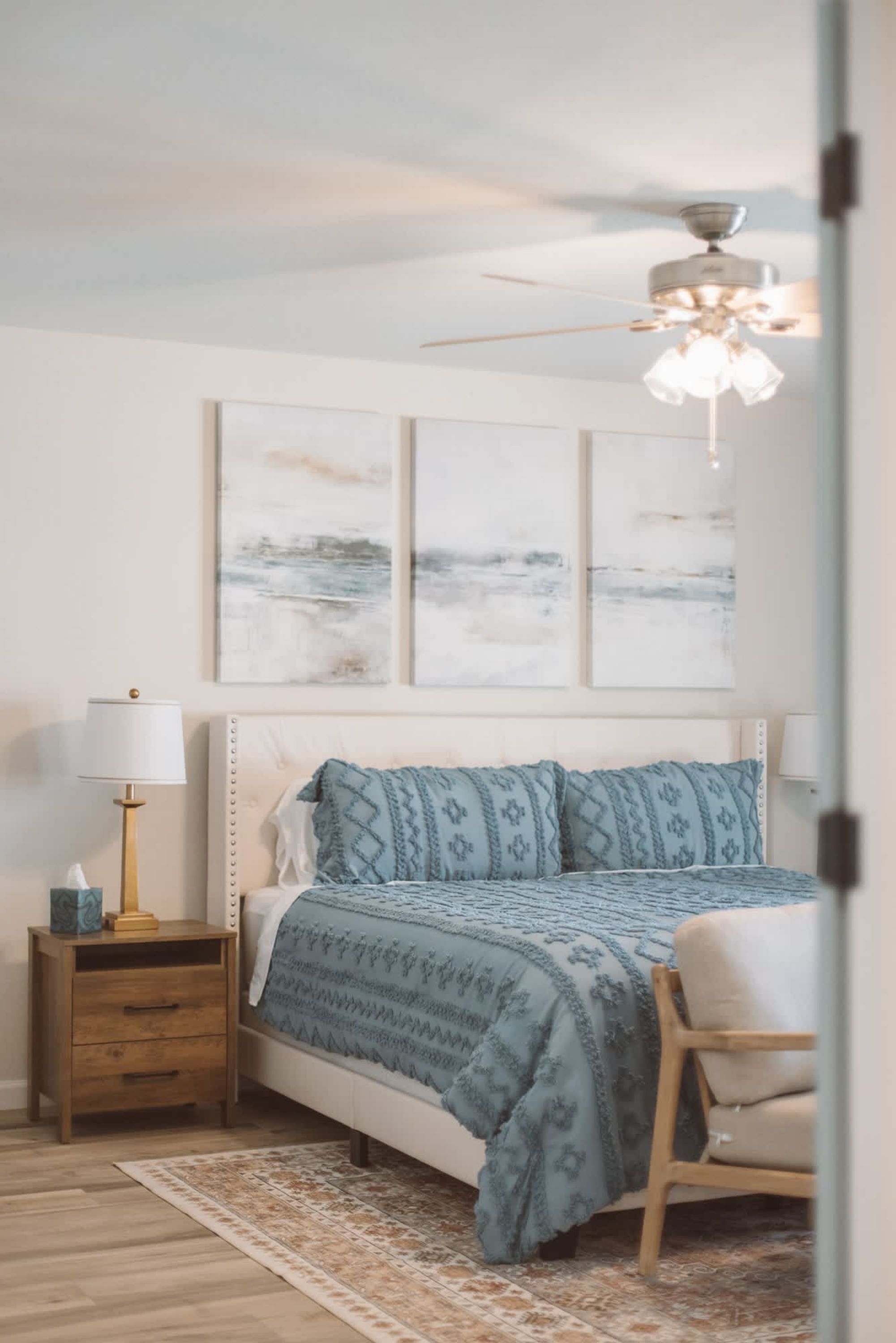 A bedroom with a bed, nightstand, lamp, chair, and ceiling fan.
