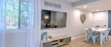 Una sala moderna con un televisor grande, un comedor con una mesa blanca y sillas azules, y un letrero en la pared que dice 'BEACH HOUSE'.