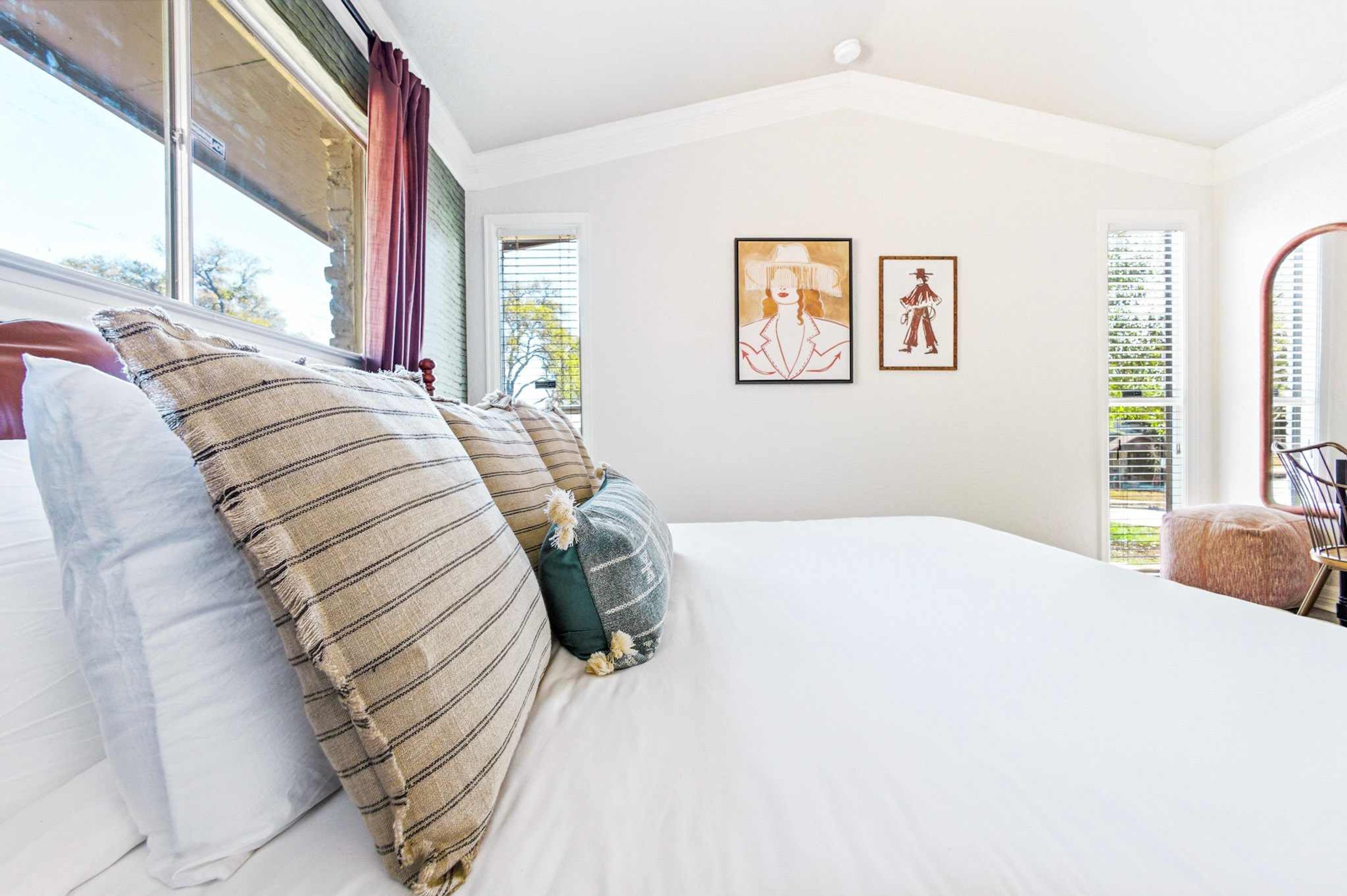 Un dormitorio con cama, almohadas, una ventana con cortinas y dos cuadros en la pared.