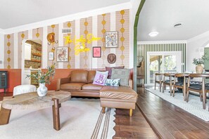 Een woonkamer met een bruine leren bank, een houten salontafel en een vloerkleed met dessin. Er is een wand met decoratieve elementen en ingekaderde foto's.