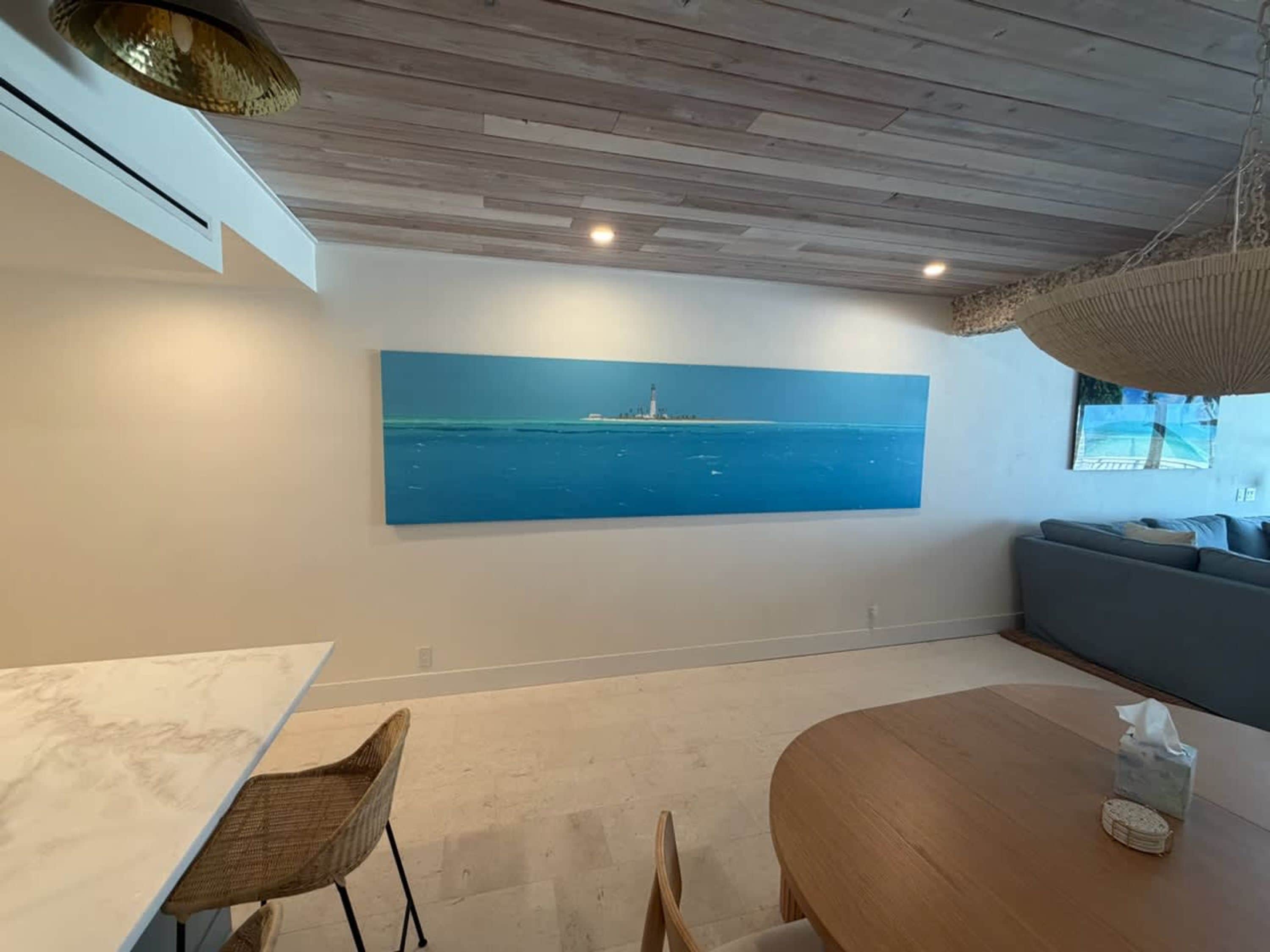 A room with a large painting of a lighthouse on the wall, a wooden ceiling, a dining table, and a sofa.
