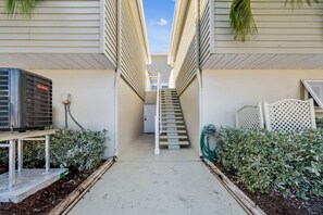 A narrow pathway leading to a staircase, flanked by walls with vertical siding and a central air conditioning unit. - Beachfront Bliss - Outdoor Pool - Sleeps 6 (Belleair Beach)