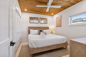 A bedroom with a wooden bed, a ceiling fan, and a window with a view of a beach. - Beachfront Bliss - Outdoor Pool - Sleeps 6 (Belleair Beach)