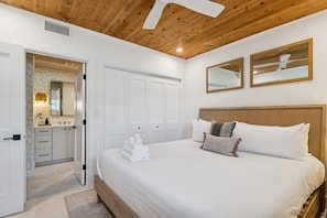 A bedroom with a wooden ceiling, white walls, a bed with white bedding, two mirrors, and a bathroom with a vanity and mirror. - Beachfront Bliss - Outdoor Pool - Sleeps 6 (Belleair Beach)