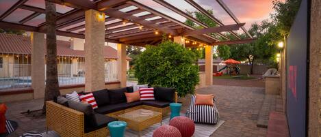 Un área de patio exterior con una zona de estar cubierta, que incluye un sofá y pufs, y vistas a una piscina al fondo.