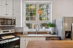Eine Küche mit weißen Schränken, einem Edelstahl-Kühlschrank, einer Mikrowelle, einem Spülbecken und einem Fenster mit Blick auf Bäume.