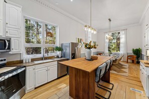 Une cuisine moderne avec des meubles blancs, un îlot central en bois, des appareils électroménagers en acier inoxydable et de grandes fenêtres offrant une vue sur les arbres.