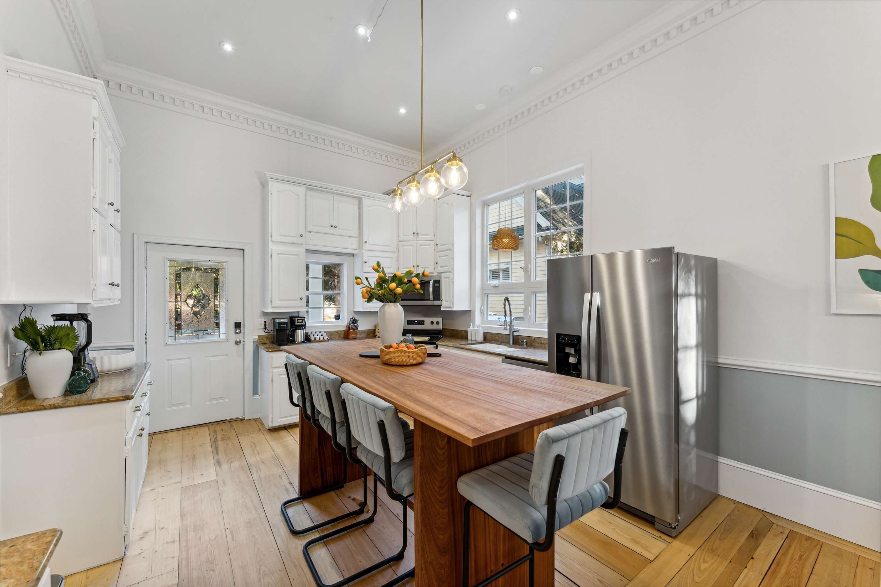 Une cuisine moderne avec des meubles blancs, un îlot central en bois doté de tabourets, un réfrigérateur en acier inoxydable et un luminaire suspendu.